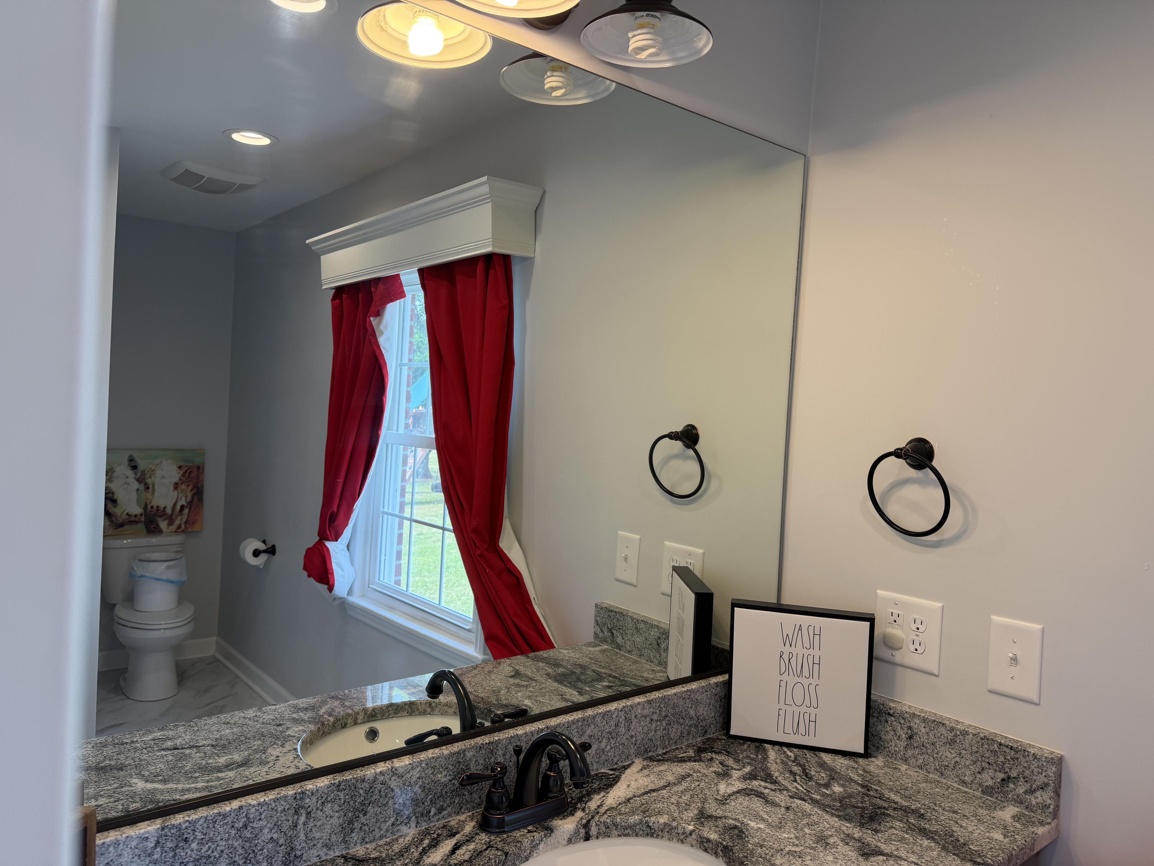 A bathroom interior with a granite countertop, sink with black fixtures, and a large mirror reflecting a window with red curtains. The counter holds a framed sign and black towel rings are mounted on the wall.